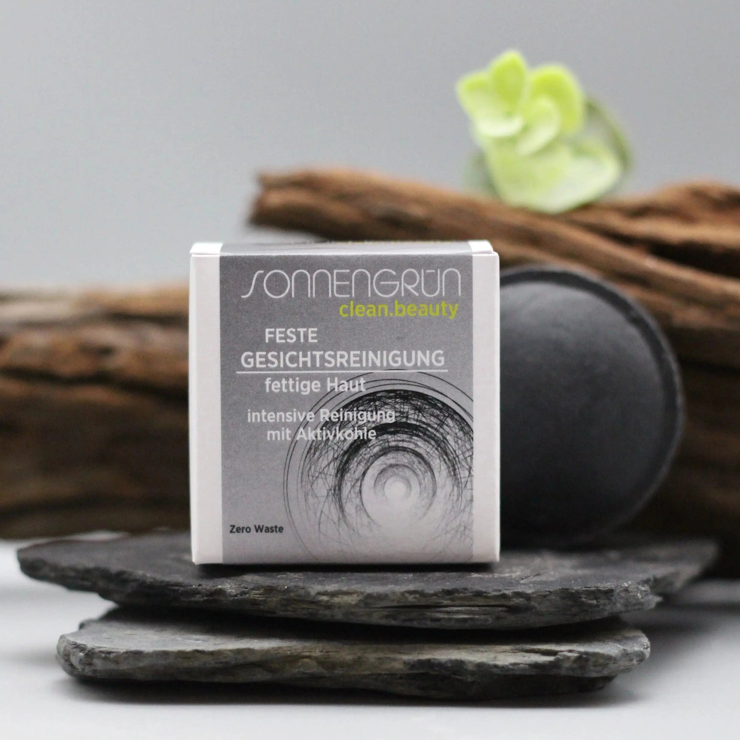 im Vordergrund des Bildes ist ein weißer, kleiner quadratischer Karton mit schwarzem Etikett zu sehen. Auf dem Etikett steht feste Gesichtsreinigung bei fettiger Haut. Im Hintergrund des Kartons sieht man eine feste, schwarze Gesichtsreinigung, da mit Aktivkohle hergestellt. Im Hintergrund sieht man Holz und ein grünes Blatt um Natürlichkeit zu vermitteln.