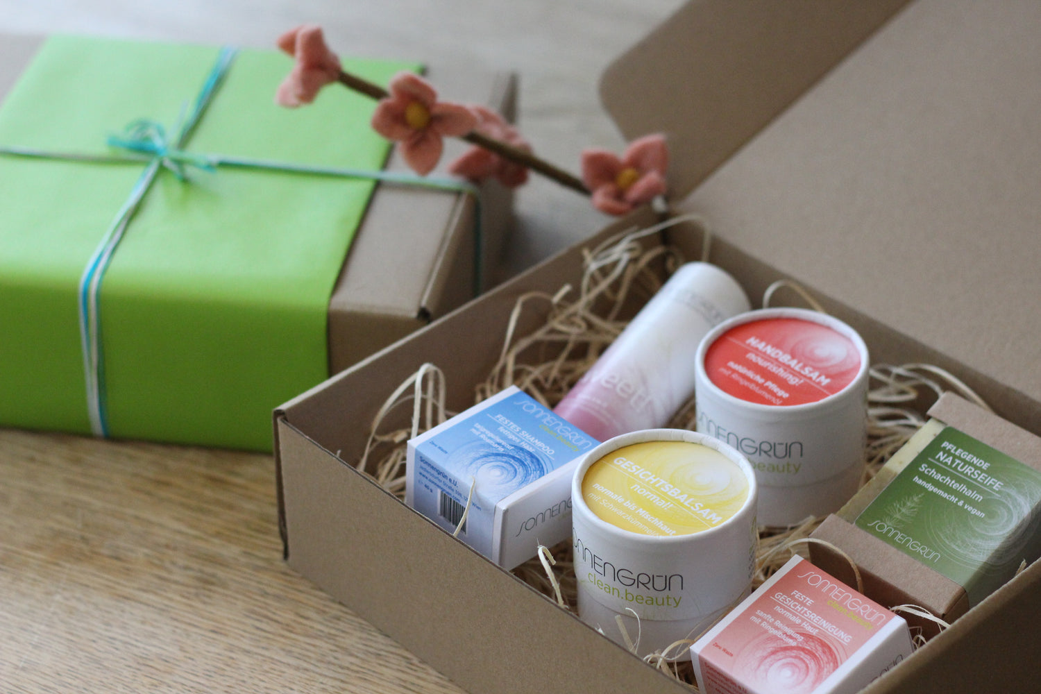 Auf dem Foto sieht man die Produkte von Sonnengrün mit ihren bunten Etiketten in einem hellbraunen Karton mit Holzwolle. Daneben sieht man ein mit grünem Geschenkpapier verpacktes Geschenk, dekoriert mit einem rosa blühendem Zweit.