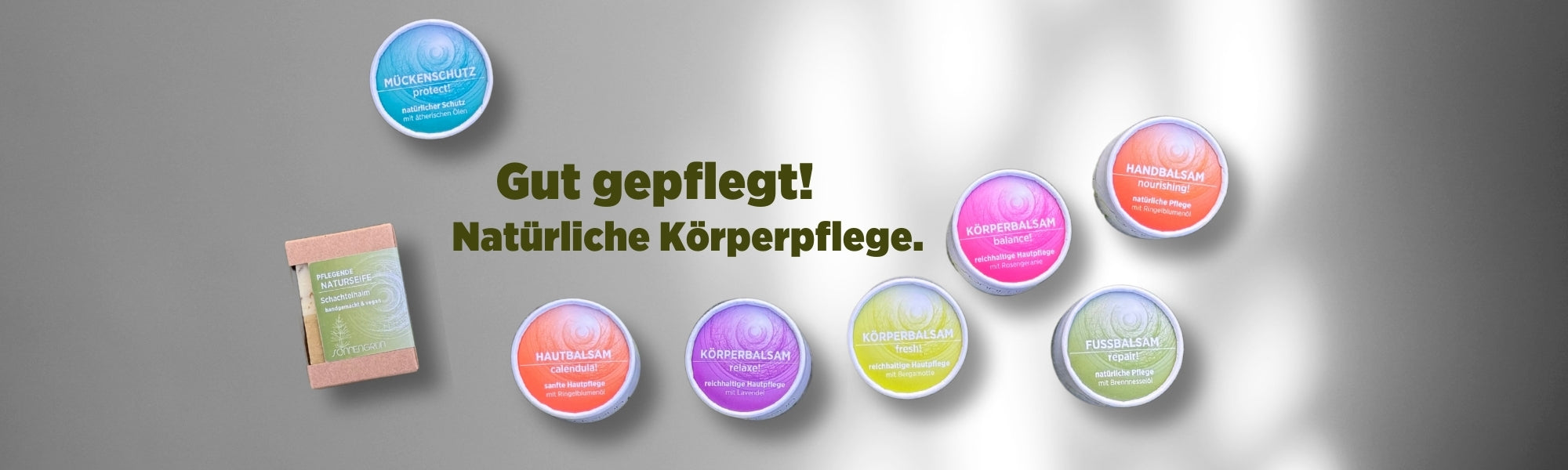 Auf dem Banner sieht man die bunten Pflegebalsame von Sonnengrün und die Naturseife mit den farbigen Etiketten in Gelb, Türkis, Lila, Orange, Grün und Rosa vor einer weißen Wand mit Schattenspiel. Mit dunkelgrüner Schrift ist zu lesen "Gut gepflegt! Natürliche Körperpflege."