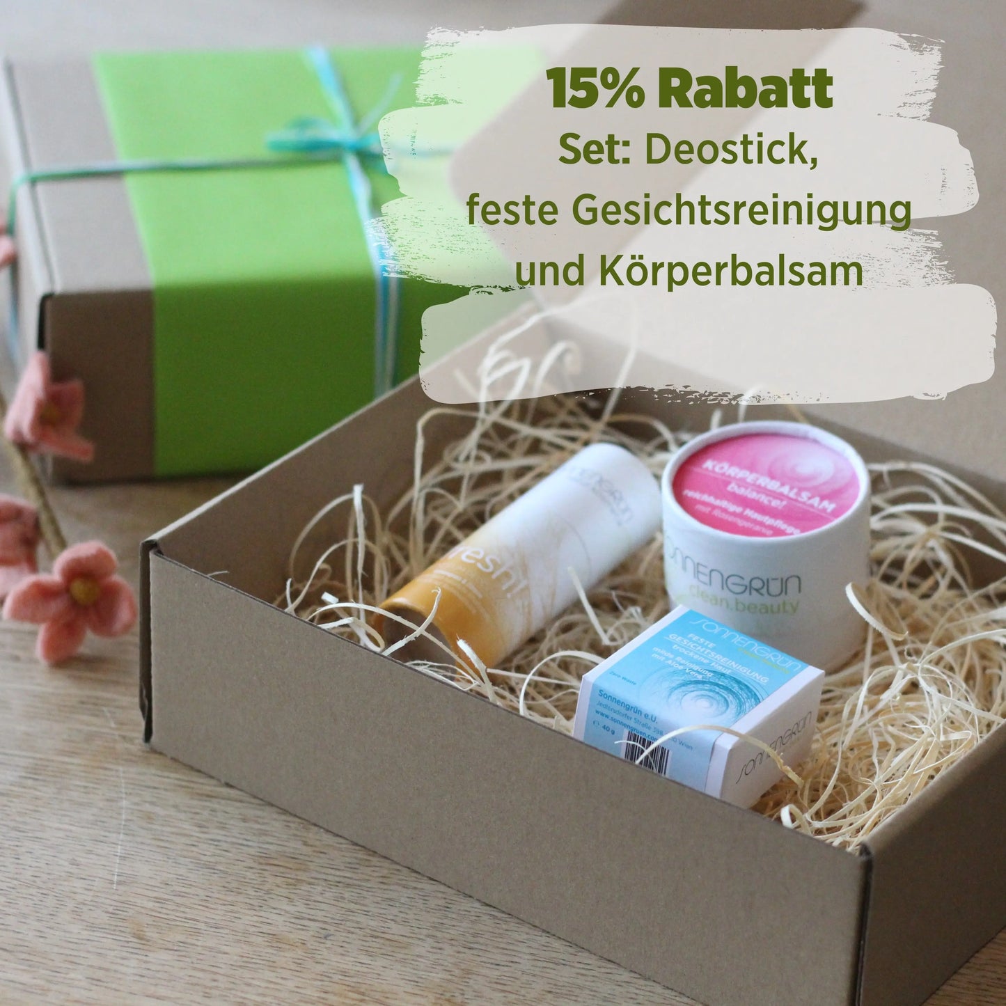 Auf dem Bild sieht man einen braunen Karton mit Holzwolle und drei Kosmetikprodukten von Sonnengrün (Deostidk, Körperbalsam und Gesichtsreinigung). Hinter der Box ist ein grün verpacktes Geschenk mit Schleife zu sehen.