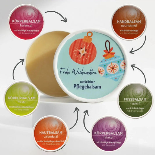 Auf dem Bild sieht man den Körperbalsam mit Weihnachtsetikett. Auf dem Etikett sind weihnachtliche Motive abgebildet. Rund um den Balsam sind die unterschiedlichen Sorten der Pflegebalsame abgebildet, Pfeile zeigen auf den feierlich gestalteten Balsam, was signalisieren soll, dass die einzelnen Balsame mit dem Weihnachtsetikett gestaltet werden können.