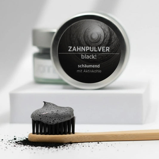 Auf dem Bild sieht man in einem Glastiegel mit Metalldeckel das schäumende Zahnpulver von Sonnengrün mit Aktivkohle. Der Tiegel ist auf kleinen Schieferplatten vor einem Hintergrund mit einem Ast aus Holz und grünen Blättern platziert. Neben dem Tiegel ist das schwarze Zahnpulver aufgehäuft.