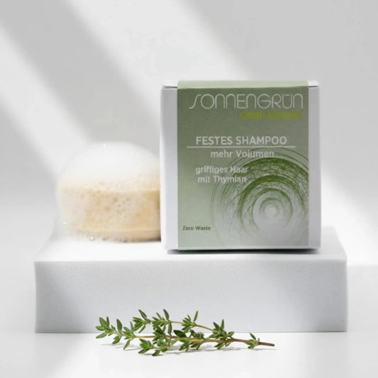 Auf dem Bild sieht man das runde feste Shampoo von Sonnengrün abgebildet. Es ist hinter der quadratischen Verpackung aus Karton mit grünem Etikett platziert.