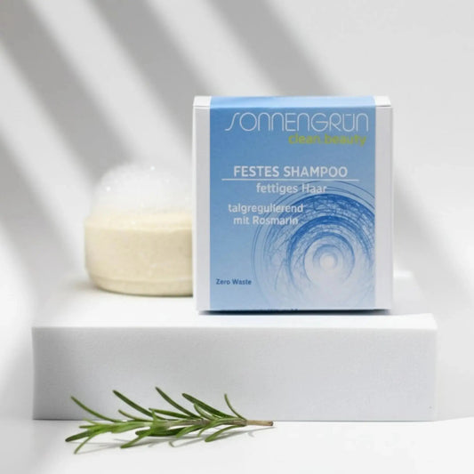 Auf dem Bild sieht man das runde feste Shampoo von Sonnengrün abgebildet. Es ist hinter der quadratischen Verpackung aus Karton mit blauem Etikett platziert. Im Hintergrund liegt ein Ast mit grünen Blättern. Das Shampoo inkl. Karton steht auf schwarzen Schieferplatten.
