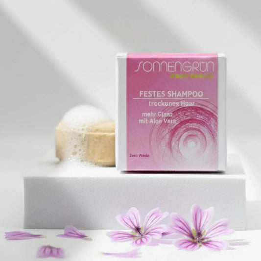 Auf dem Bild sieht man das runde feste Shampoo von Sonnengrün abgebildet. Es ist hinter der quadratischen Verpackung aus Karton mit rosa Etikett platziert.