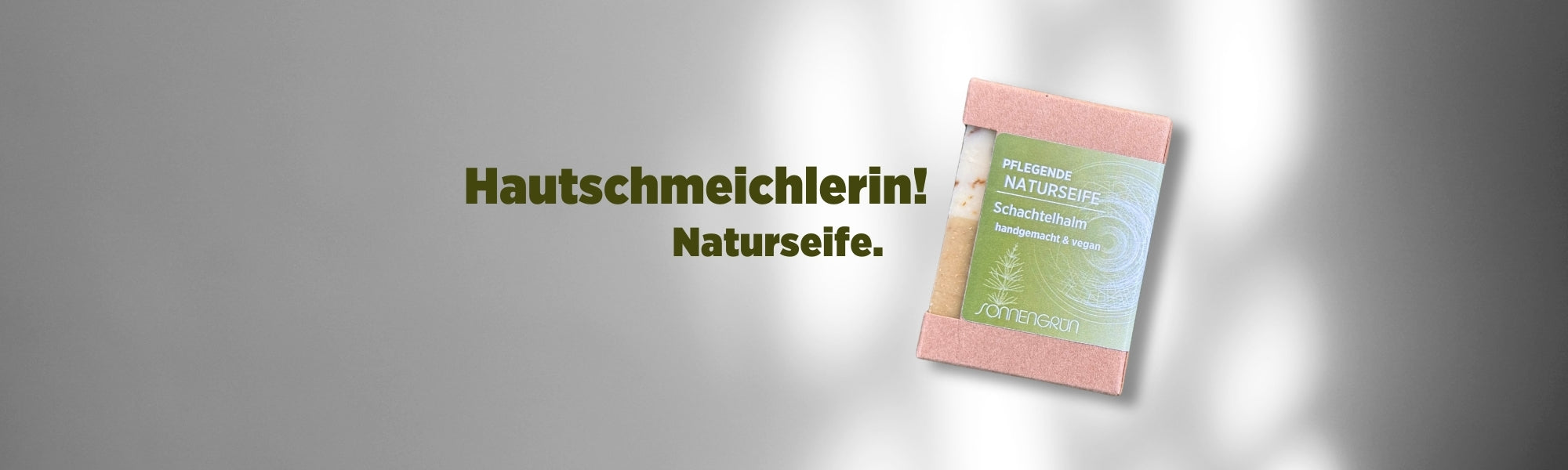 Auf dem Banner sieht man die Naturseife von Sonnengrün in Karton verpackt mit grünem Etikett vor einer weißen Wand mit Schattenspiel. Mit dunkelgrüner Schrift ist zu lesen "Hautschmeichlerin! Naturseife."