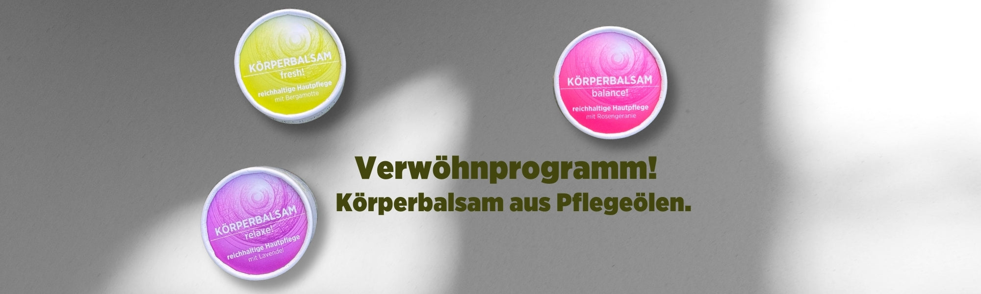 Auf dem Banner sieht man die bunten Körpercremen von Sonnengrün in den farbigen Rundkartonagen Gelb, Rosa und Lila vor einer weißen Wand mit Schattenspiel. Mit dunkelgrüner Schrift ist zu lesen "Verwöhnprogramm! Körperbalsam aus Pflegeölen."