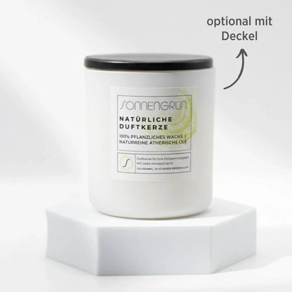 Auf diesem Foto sieht man die Duftkerze von Sonnengrün in einem weißem Glas auf einem weißen Podest stehen. Die Naturkerze ist mit einem Holzdeckel verschlossen, was mit dunkler Schrift gekennzeichnet ist ("optional mit Decke").