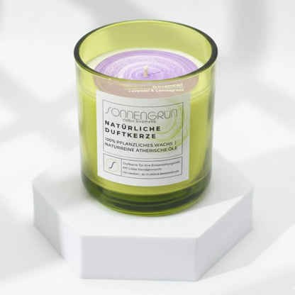 Auf diesem Foto sieht man die Duftkerze von Sonnengrün in einem grünem Glas auf einem weißen Podest stehen.