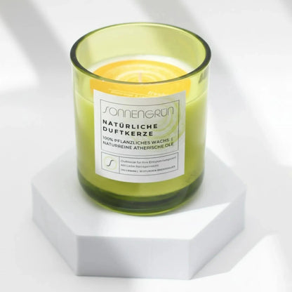 Auf diesem Foto sieht man die Duftkerze von Sonnengrün in einem grünen Glas auf einem weißen Podest stehen.