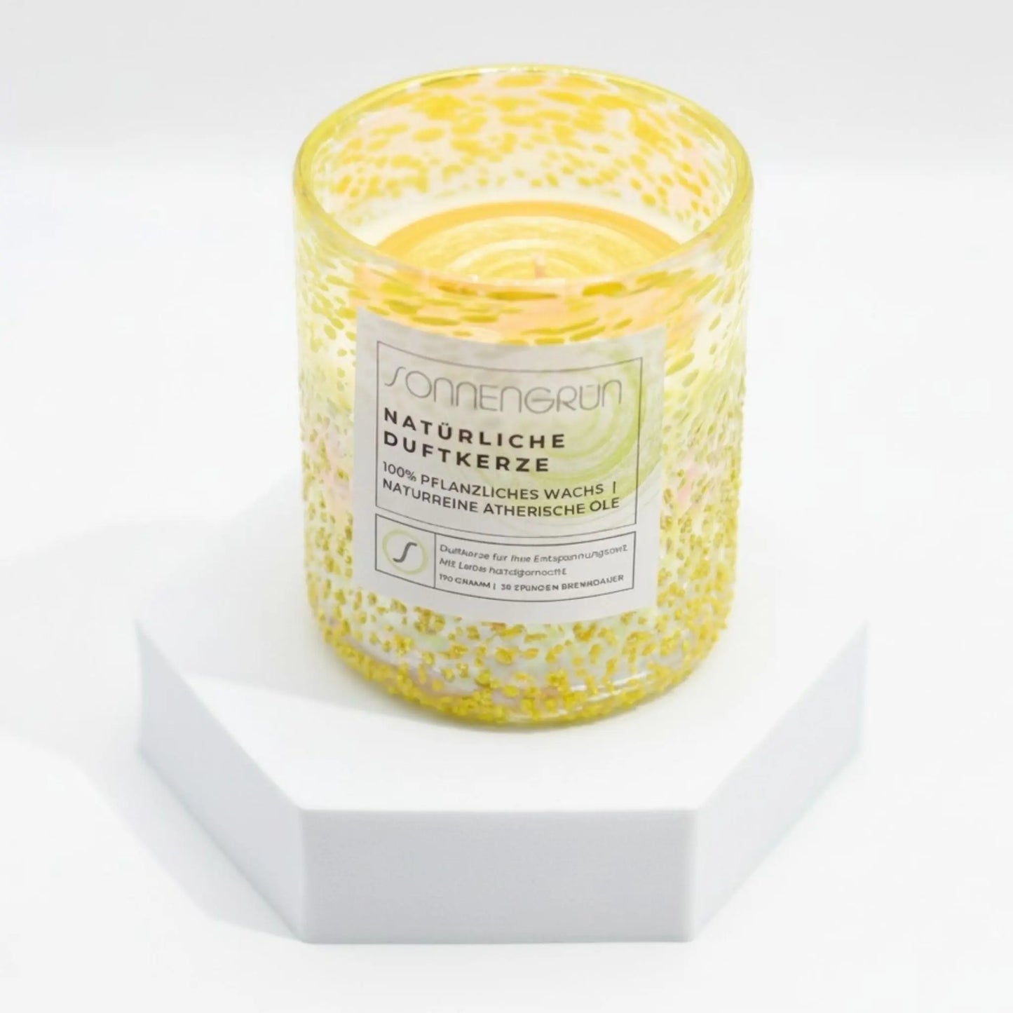 Auf diesem Foto sieht man die Duftkerze von Sonnengrün in einem gelben Glas auf einem weißen Podest stehen.