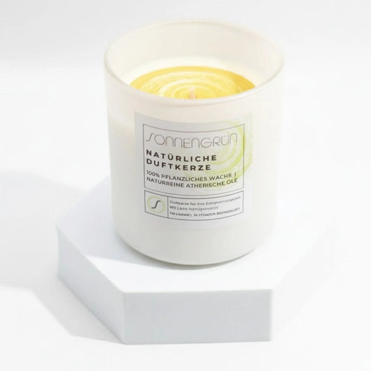 Auf diesem Foto sieht man die Duftkerze von Sonnengrün in einem weißen Glas auf einem weißen Podest stehen.