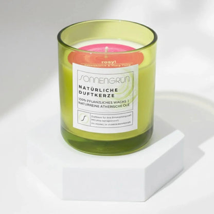 Auf diesem Foto sieht man die Duftkerze von Sonnengrün in einem grünen Glas auf einem weißen Podest stehen.