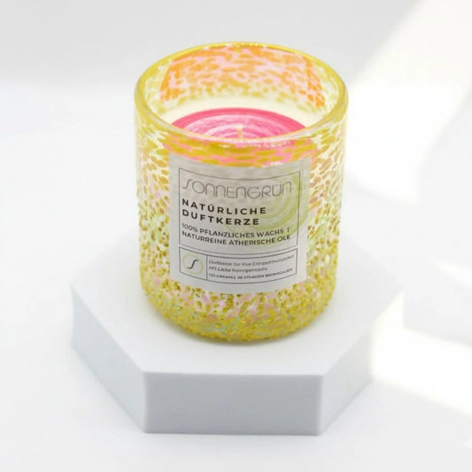 Auf diesem Foto sieht man die Duftkerze von Sonnengrün in einem gelben Glas auf einem weißen Podest stehen.