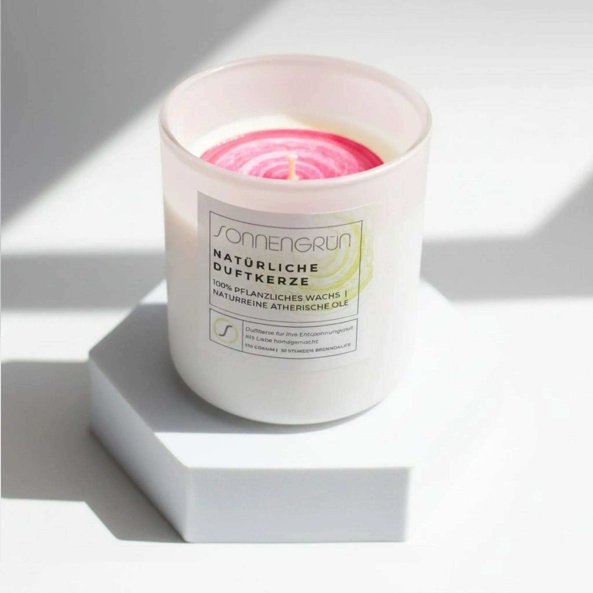 Auf diesem Foto sieht man die Duftkerze von Sonnengrün in einem weißen Glas auf einem weißen Podest stehen.