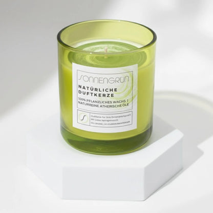 Auf diesem Foto sieht man die Duftkerze von Sonnengrün in einem grünem Glas auf einem weißen Podest stehen.