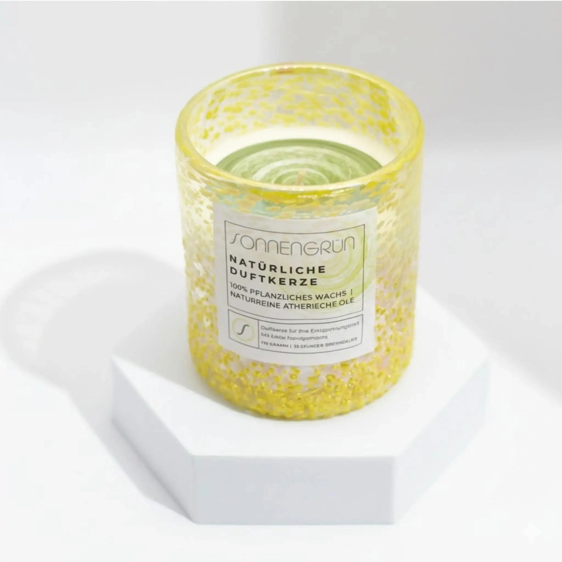 Auf diesem Foto sieht man die Duftkerze von Sonnengrün in einem grünem Glas auf einem weißen Podest stehen.