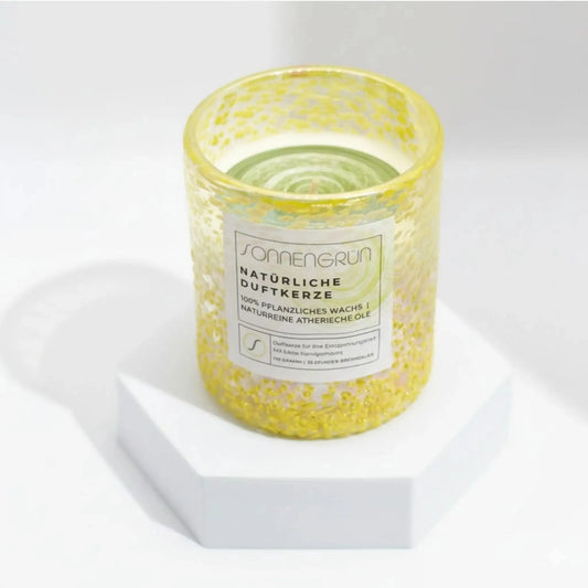 Auf diesem Foto sieht man die Duftkerze von Sonnengrün in einem grünem Glas auf einem weißen Podest stehen.
