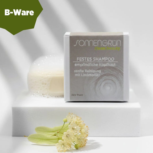 Auf dem Bild sieht man das runde feste Shampoo von Sonnengrün abgebildet. Es ist hinter der quadratischen Verpackung aus Karton mit beigen Etikett platziert. B-Ware.
