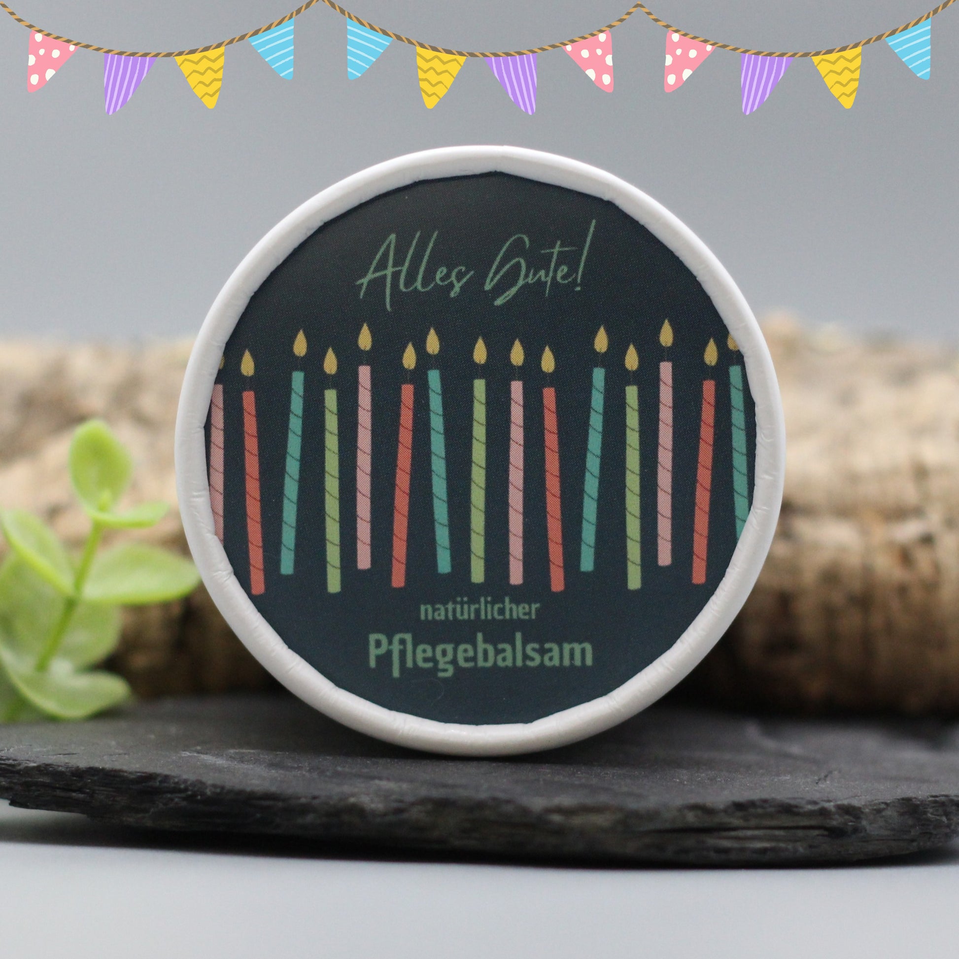 Auf dem Bild sieht man den Gesichtsbalsam mit einem Etikett, gestaltet speziell für Geburtstage. Auf dem Etikett sich man bunte Kerzen und die Aufschrift "Alles Gute!" und "natürlicher Pflegebalsam". Das Etikett macht den Gesichtsbalsam zum perfekten Geburtstagsgeschenk.