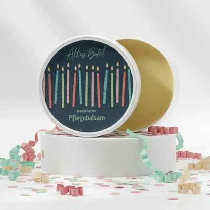 Auf dem Bild sieht man den Gesichtsbalsam mit einem Etikett, gestaltet speziell für Geburtstage. Auf dem Etikett sich man bunte Kerzen und die Aufschrift "Alles Gute!" und "natürlicher Pflegebalsam". Das Etikett macht den Gesichtsbalsam zum perfekten Geburtstagsgeschenk.