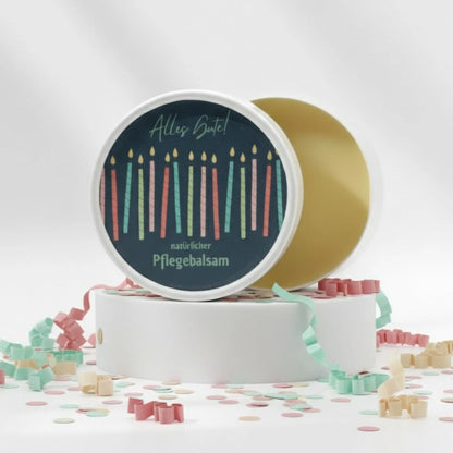 Auf dem Bild sieht man den Pflegebalsam mit einem Etikett, gestaltet speziell für Geburtstage. Auf dem Etikett sich man bunte Kerzen und die Aufschrift "Alles Gute!" und "natürlicher Pflegebalsam". Das Etikett macht den Körperbalsam zum perfekten Geburtstagsgeschenk.