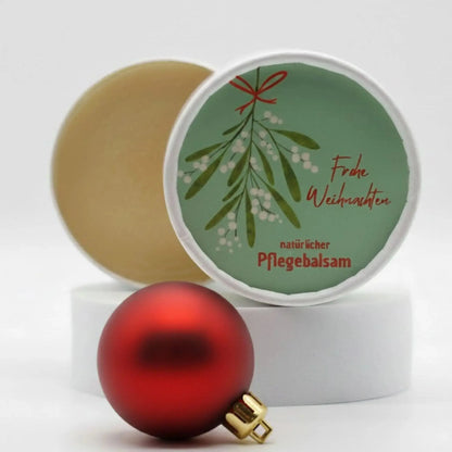 Auf dem Bild sieht man den Pflegebalsam mit einem Etikett, gestaltet speziell für Weihnachten. Auf dem Etikett sieht man ein weihnachtliches Motiv und die Aufschrift "Frohe Weihnachten!" und "natürlicher Pflegebalsam". Das Etikett macht den Pflegebalsam zum perfekten Weihnachtsgeschenk.
