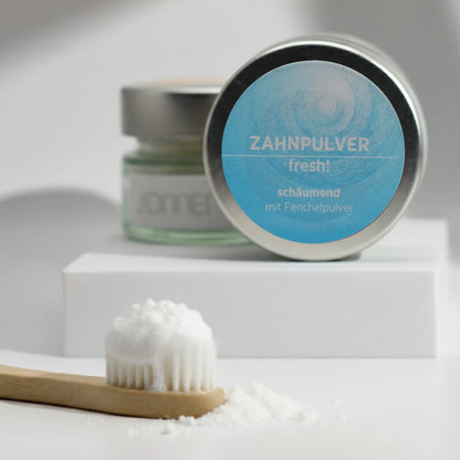 Auf dem Bild sieht man das schäumende Zahnpulver von Sonnengrün im Glastiegel und Metalldecke auf zwei kleinen Schieferplatten platziert. Im Hintergrund sieht man einen Ast und grüne Blätter, die das Bild natürlich erscheinen lassen.