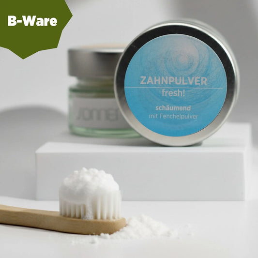 Auf dem Bild sieht man das schäumende Zahnpulver von Sonnengrün im Glastiegel und Metalldecke auf zwei kleinen Schieferplatten platziert. Im Hintergrund sieht man einen Ast und grüne Blätter, die das Bild natürlich erscheinen lassen. B-Ware.