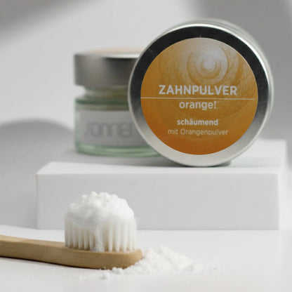Auf dem Bild sieht man das schäumende Zahnpulver mit Orangengeschmack von Sonnengrün in einem Glastiegel mit Metalldeckel auf kleinen schwarzen Schieferplatten vor grauem Hintergrund platziert. Im Hintergrund sieht man einen braunen Ast und ein paar Blätter, die das Bild natürlich erscheinen lassen.