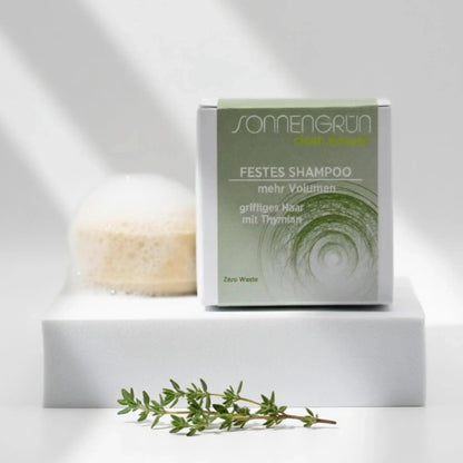 Auf dem Bild sieht man das runde feste Shampoo von Sonnengrün abgebildet. Es ist hinter der quadratischen Verpackung aus Karton mit grünem Etikett platziert.