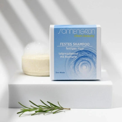 Auf dem Bild sieht man das runde feste Shampoo von Sonnengrün abgebildet. Es ist hinter der quadratischen Verpackung aus Karton mit blauem Etikett platziert. Im Hintergrund liegt ein Ast mit grünen Blättern. Das Shampoo inkl. Karton steht auf schwarzen Schieferplatten.