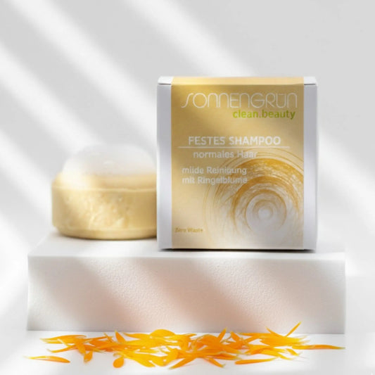 Auf dem Bild sieht man das runde feste Shampoo von Sonnengrün abgebildet. Es ist hinter der quadratischen Verpackung aus Karton mit gelben Etikett platziert. 